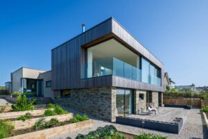 Modern two-story house with a wooden and stone exterior, large glass windows, and double glazed doors leading to a patio. It's surrounded by a garden featuring raised wooden planters under a clear blue sky.