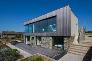 A modern two-story house with large glass windows, double glazed doors, stone detailing, and an outdoor patio, set against a clear blue sky.