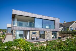 Modern two-story house with large glass windows and stone accents, featuring sleek double glazed doors, surrounded by greenery under a clear blue sky.