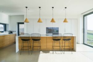 Modern kitchen with four wire-frame bar stools at a wooden island, pendant lights above, and large windows on the right. White cabinets and built-in oven in the background complete this sleek space, while double glazed doors provide both style and energy efficiency.