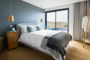 A modern bedroom features a large bed, two wooden side tables, and hanging lamps. Double glazed sliding glass doors lead to a balcony. Neutral tones and simple decor create a calm atmosphere.