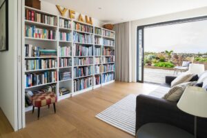 A bright room with a large bookshelf, a gray sofa, and a small stool. Double glazed glass doors open to an outdoor area. Wooden floor and white walls create a clean, modern look. "YEAH" letters on top of the shelf add a playful touch.