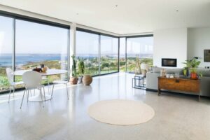 Modern living room with large windows and double glazed doors offering a coastal view. Features a round table with chairs, a sofa, plants, and a wooden chest on a sleek floor.