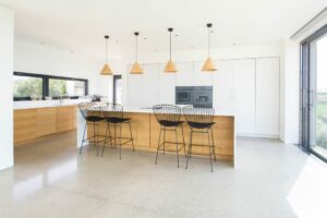 Modern kitchen with a large island, wooden cabinetry, black wire bar stools, pendant lights, and floor-to-ceiling windows featuring elegant double glazed doors.