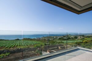 With double glazed doors open wide, enjoy a breathtaking view from the balcony overlooking a coastal landscape teeming with fields, distant houses, and the serene sea under a clear blue sky.
