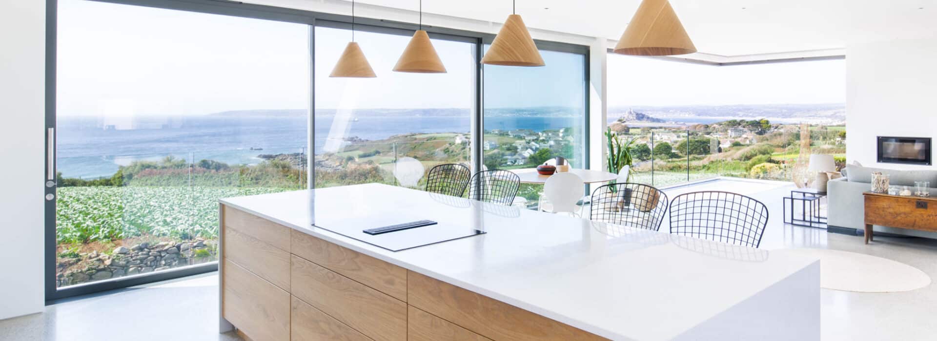 Modern open-plan kitchen with a large island, hanging lights, and floor-to-ceiling windows offering a panoramic ocean view. Double glazed doors enhance the dining and living areas visible in the background.