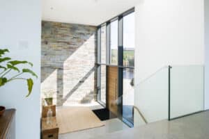 Modern interior entryway with large windows, stone accent wall, and glass railing. Corner sliding doors enhance the space, while a small plant and wooden bench allow natural light to brighten the area.