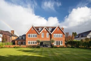 Large two-story brick house with white trim, dormer windows, and elegant balustrades. A well-kept lawn enhances its charm, while a rainbow arcs across the partly cloudy sky.