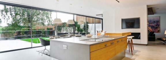 Modern kitchen with a large island, bar stools, pendant lights, and floor-to-ceiling windows complemented by sleek sliding aluminium doors that open to a garden view.