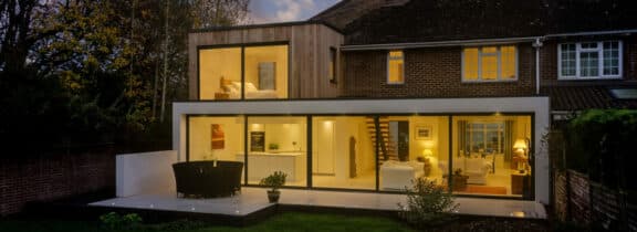 Two-story modern house extension with large glass windows and sliding aluminium doors, a wooden upper facade, and a lit interior showcasing a living area and kitchen, viewed at dusk.