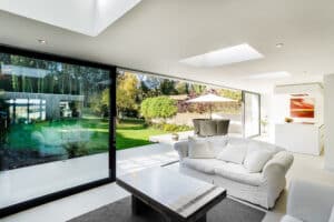 Modern living room with white furniture and a large window featuring sliding aluminium doors, overlooking a green garden. A dining table is visible outside on the patio.