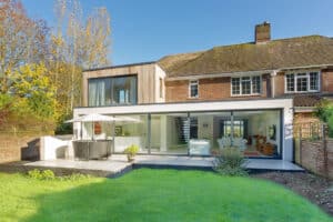 A modern house extension featuring sliding aluminium doors, a patio with furniture, and a lush green lawn in front.