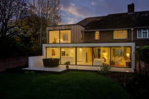 Modern house with large glass windows and sliding aluminum doors, illuminated interior, and a two-story extension. Cozy living room, open kitchen, and dining area visible. Night scene with a well-lit garden.