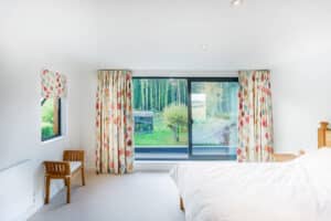 The bright bedroom features white walls and a large window adorned with floral curtains, offering a view of the garden. Sliding aluminium doors add a modern touch, complementing the cozy bed and chairs in the space.