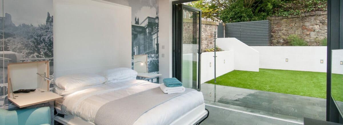 Modern bedroom with sleek aluminium bifold doors opening to a small outdoor yard. The room features a bed, bedside table, and a wall artwork showcasing a cityscape.