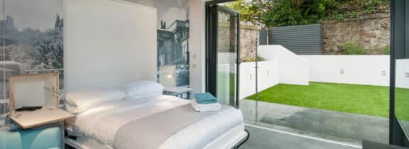 Modern bedroom with sleek aluminium bifold doors opening to a small outdoor yard. The room features a bed, bedside table, and a wall artwork showcasing a cityscape.