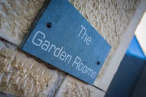 A sleek sign reading "The Garden Rooms" is mounted on a textured stone wall, seamlessly complemented by modern aluminium bifolds that invite natural light and elegance into the space.