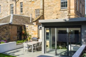 A modern patio features a dining table, chairs, and a barbecue grill, seamlessly connected to the contemporary extension’s sleek aluminium bifolds while complementing the charm of an adjacent older stone building.