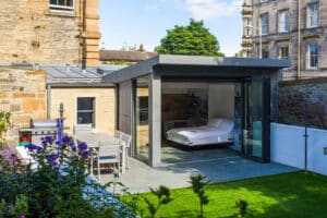 Modern patio with an aluminium bifold extension, revealing a bed indoors through the elegant sliding glass door. The patio includes a dining table and chairs, perfectly nestled within a lush garden setting.