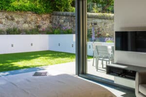 The bedroom offers a serene view with a cozy bed, wall-mounted TV, and sleek aluminium bifolds opening to a patio. Outside, the grassy lawn invites relaxation, complemented by a table, chairs, and a charming stone wall backdrop.