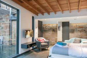 Modern bedroom with exposed wooden beams, a bed, a wall-mounted TV, and a UK flag-themed chair. Stone walls and sleek aluminium bifolds replace the sliding glass door, leading to an inviting outdoor area.