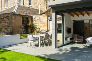 A modern patio features a dining table and chairs beside sleek aluminium bifolds, opening to a stylish interior. An older brick building is in the background, with a mix of sun and shade present.