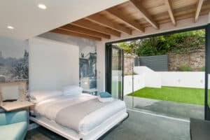 A modern bedroom featuring a fold-down bed, open aluminium bifolds leading to a small garden with artificial grass, and a wooden beamed ceiling.