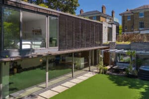 Modern two-story house with large glass windows and a sleek glass sliding door, featuring a visible bedroom and living area. The landscaped lawn offers outdoor seating under a pergola, with another house visible in the background.