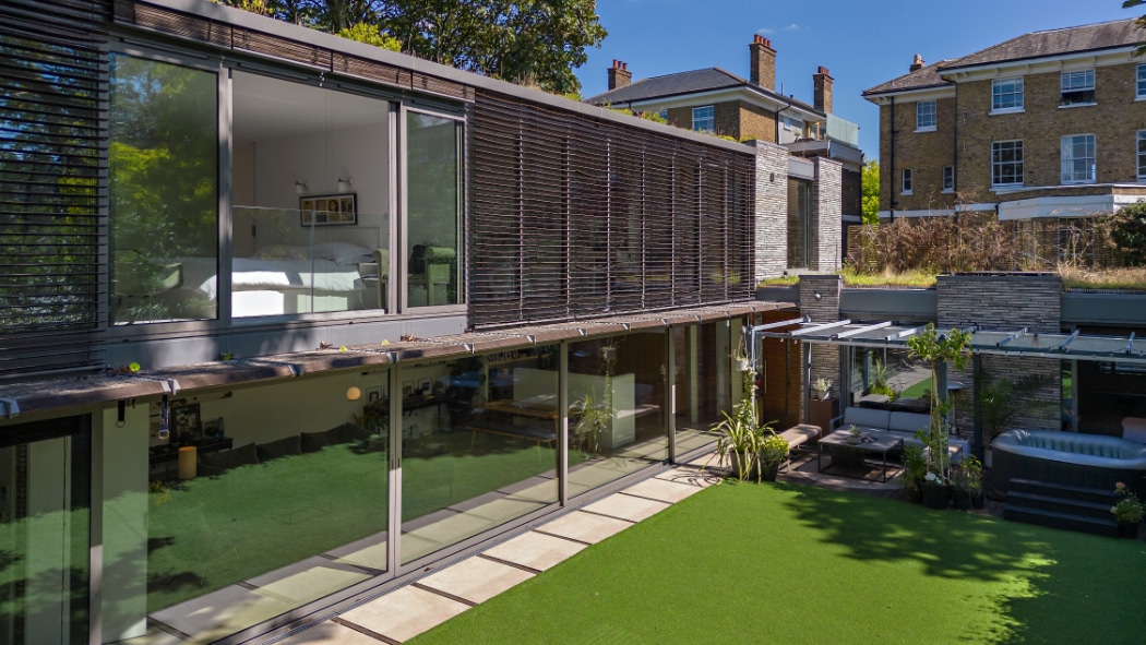 Modern two-story house with large glass windows and a sleek glass sliding door, featuring a visible bedroom and living area. The landscaped lawn offers outdoor seating under a pergola, with another house visible in the background.