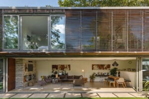 Modern two-story house with large glass windows and a sleek glass sliding door, an open-plan living area, outdoor seating, and contemporary furnishings visible inside.