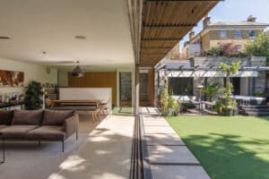 Modern open-plan living space seamlessly connects to a backyard with artificial grass, patio seating, and lush greenery, divided by a large glass sliding door.