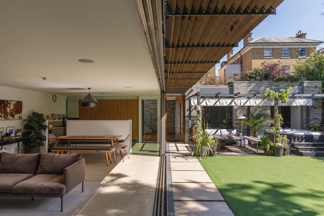 Modern open-plan living room seamlessly connects through a sleek glass sliding door to a covered outdoor patio and artificial grass yard, featuring contemporary furniture and greenery.