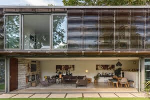Modern two-story house with large glass windows and horizontal wooden louvers; a glass sliding door opens to the open-plan living and dining area visible on the ground floor.