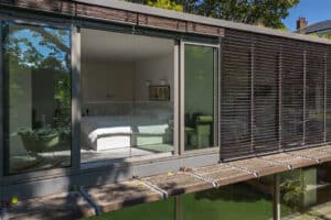 A modern bedroom with a white bed is visible through a sleek glass sliding door, featuring horizontal slatted shades outside and the reflection of trees in the window.