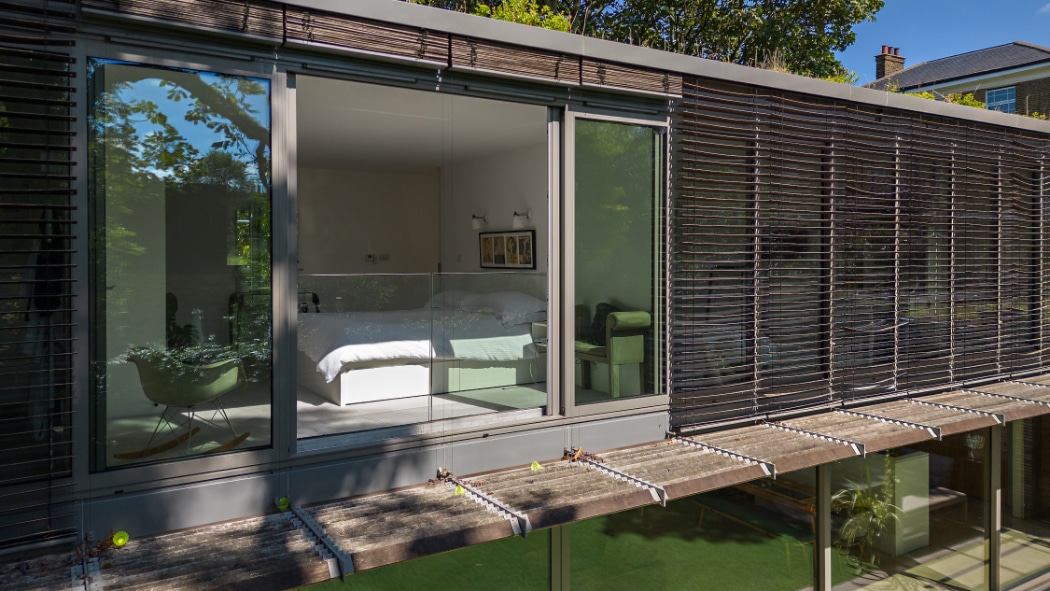 A modern bedroom with a white bed is visible through a sleek glass sliding door, featuring horizontal slatted shades outside and the reflection of trees in the window.