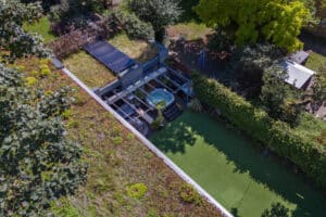 Aerial view of a modern glass-walled structure with a green roof and a glass sliding door, adjacent to a lawn area, surrounded by trees and hedges.
