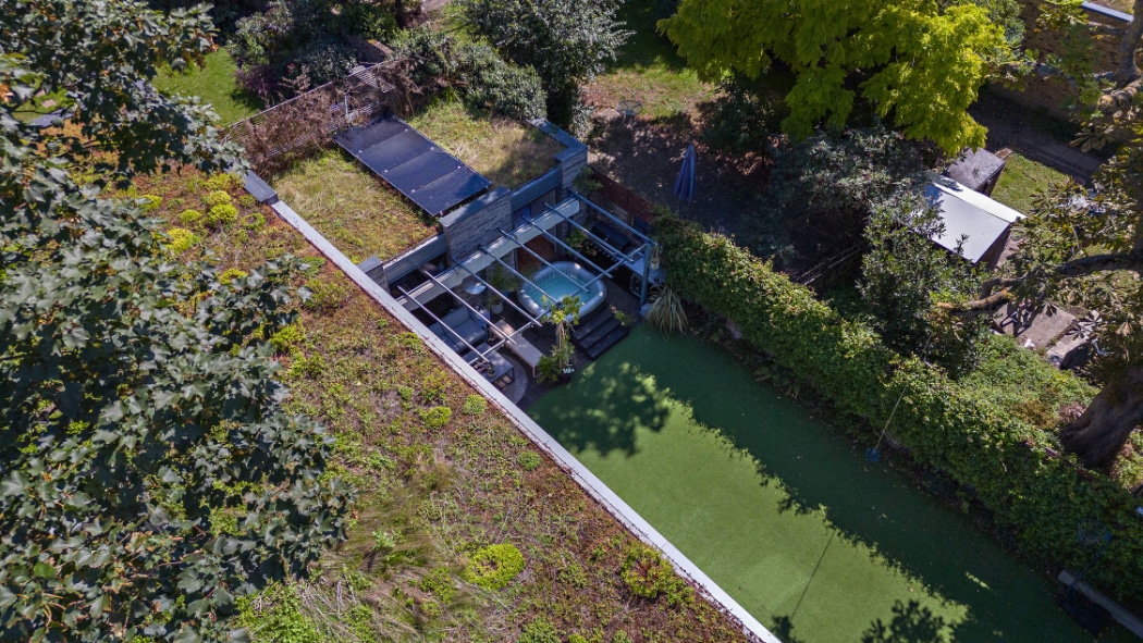Aerial view of a modern glass-walled structure with a green roof and a glass sliding door, adjacent to a lawn area, surrounded by trees and hedges.