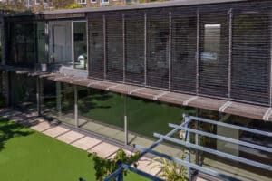 Modern building with large glass windows, a sleek glass sliding door, and horizontal external blinds overlooks a lawn and paved area under bright daylight.