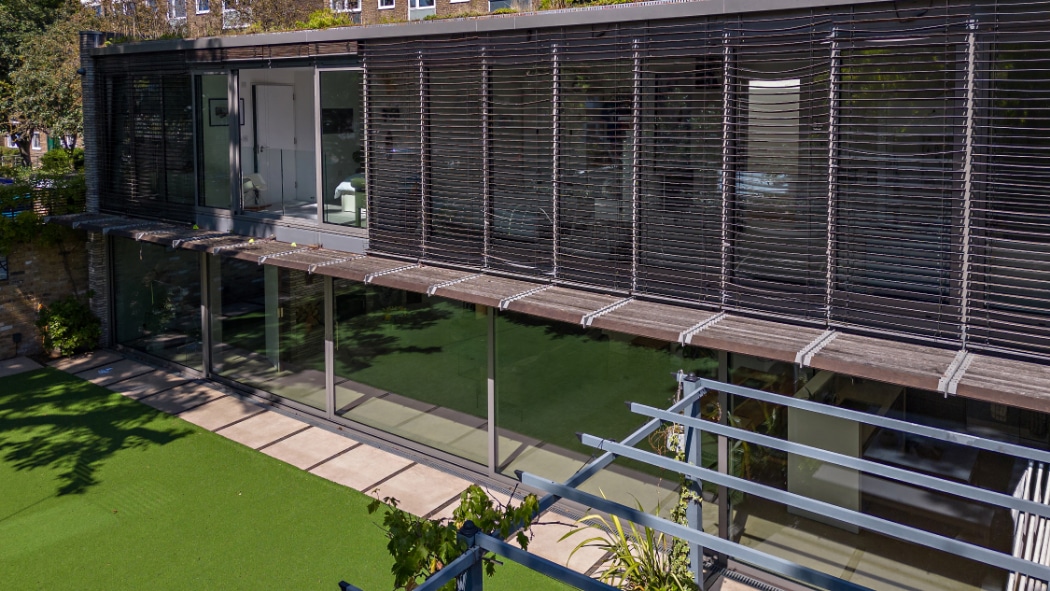 Modern building with large glass windows, a sleek glass sliding door, and horizontal external blinds overlooks a lawn and paved area under bright daylight.