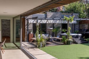 A modern patio with a glass sliding door opens to an outdoor seating area, potted plants, a jacuzzi, and a grill, all surrounded by a privacy wall and lush greenery.