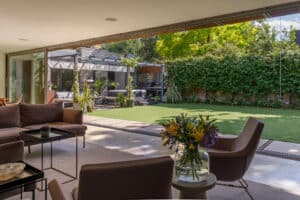 A modern living room with a glass sliding door opens to a spacious backyard featuring artificial grass, patio seating, lush plants, and outdoor furniture on a sunny day.