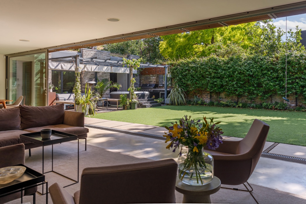 A modern living room with a glass sliding door opens to a spacious backyard featuring artificial grass, patio seating, lush plants, and outdoor furniture on a sunny day.