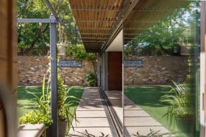Modern garden with artificial grass, potted plants, and a brick wall, reflected in large glass windows and a sleek glass sliding door beneath a wooden pergola.