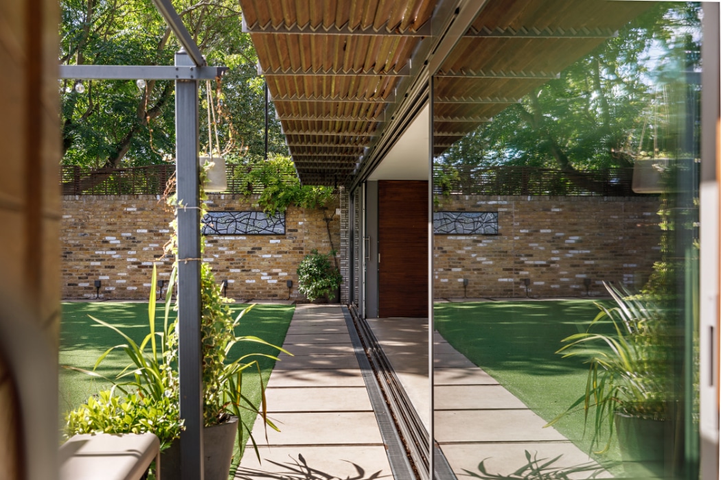 Modern garden with artificial grass, potted plants, and a brick wall, reflected in large glass windows and a sleek glass sliding door beneath a wooden pergola.