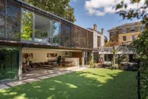Modern house with expansive glass sliding door opens onto a green artificial lawn; outdoor seating area and a traditional brick house are visible in the background under a blue sky.