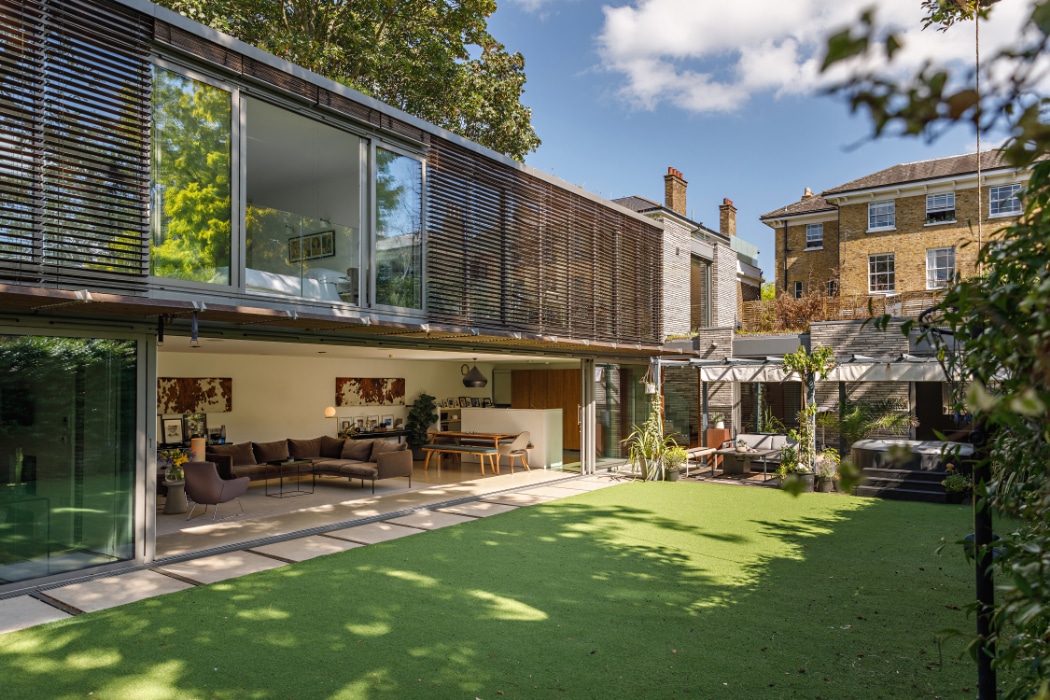 Modern house with expansive glass sliding door opens onto a green artificial lawn; outdoor seating area and a traditional brick house are visible in the background under a blue sky.