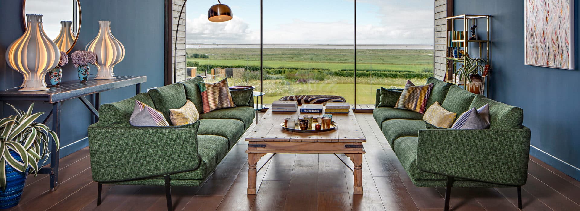 A modern living room features green sofas, a wooden coffee table, and decorative lamps. Slimline sliding doors lead to large windows, revealing a grassy landscape with water in the distance.