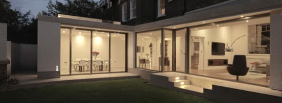 A modern glass-encased house extension showcases a well-lit interior, with sliding aluminium doors that open from the dining area and living room onto a neatly maintained lawn.