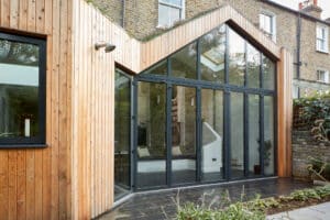 Modern house extension featuring wood paneling and large glass doors, complemented by a striking glazed gable end.