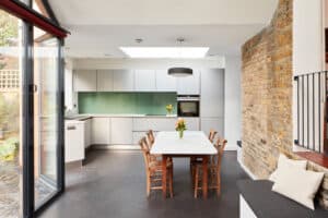 Modern kitchen with white cabinets, green backsplash, and a skylight. A wooden table with four chairs is in the center. Exposed brick wall to the right and large sliding glass doors leading to a glazed gable end on the left, offering plenty of natural light.